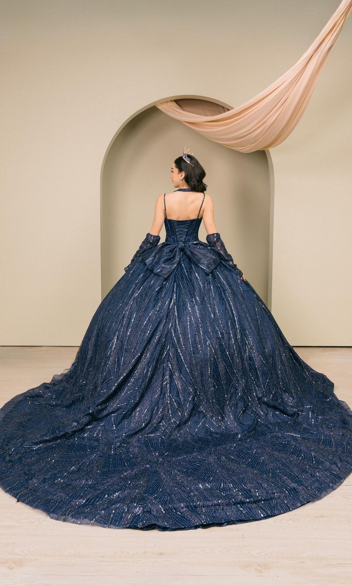 A woman stands facing an archway, wearing the Quinceanera Dress 8510JQ by Cinderella Coutureโa dark blue, sequin-embellished ball gown featuring a long train and off-shoulder sleeves. Draped fabric above the arch adds a touch of Cinderella Couture elegance to the scene.